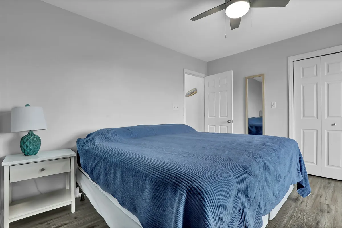 A serene bedroom with a blue bedspread, white nightstand, decorative lamp, and ceiling fan. The room exudes calm, perfect for unwinding after a day at the beach, and showcases a layout ideal for guests or family.