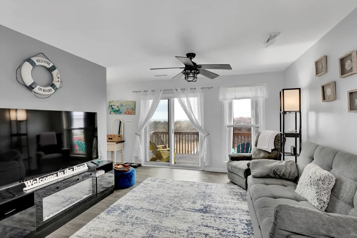 A cozy living room featuring a large grey sofa (which could double as a sleeper), a big television, and a set of large windows letting in plenty of sunlight. Decorative wall art and a relaxed seating arrangement set a welcoming mood, while the windows connect the indoors to the ocean scenery outside.