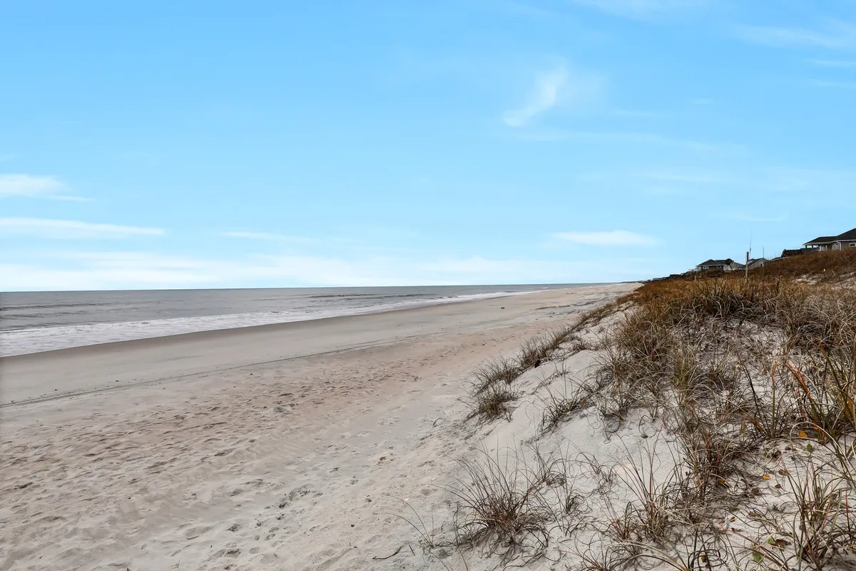 A tranquil stretch of sandy beach with gentle waves, dune grass, and houses visible in the background. The vista highlights the natural beauty and open space available for group activities like sunbathing, beachcombing, and water sports.