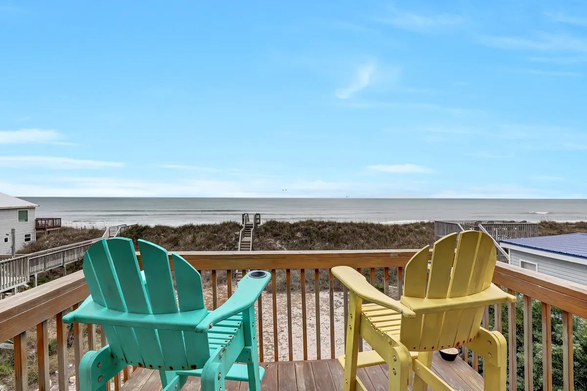 The image features a serene beachside setting at North Topsail Beach, with bright turquoise and yellow Adirondack chairs arranged on a deck overlooking the ocean. A private boardwalk leads directly from the house over the dunes to the sandy shore, and the expansive sea view invites relaxation and connection with nature.