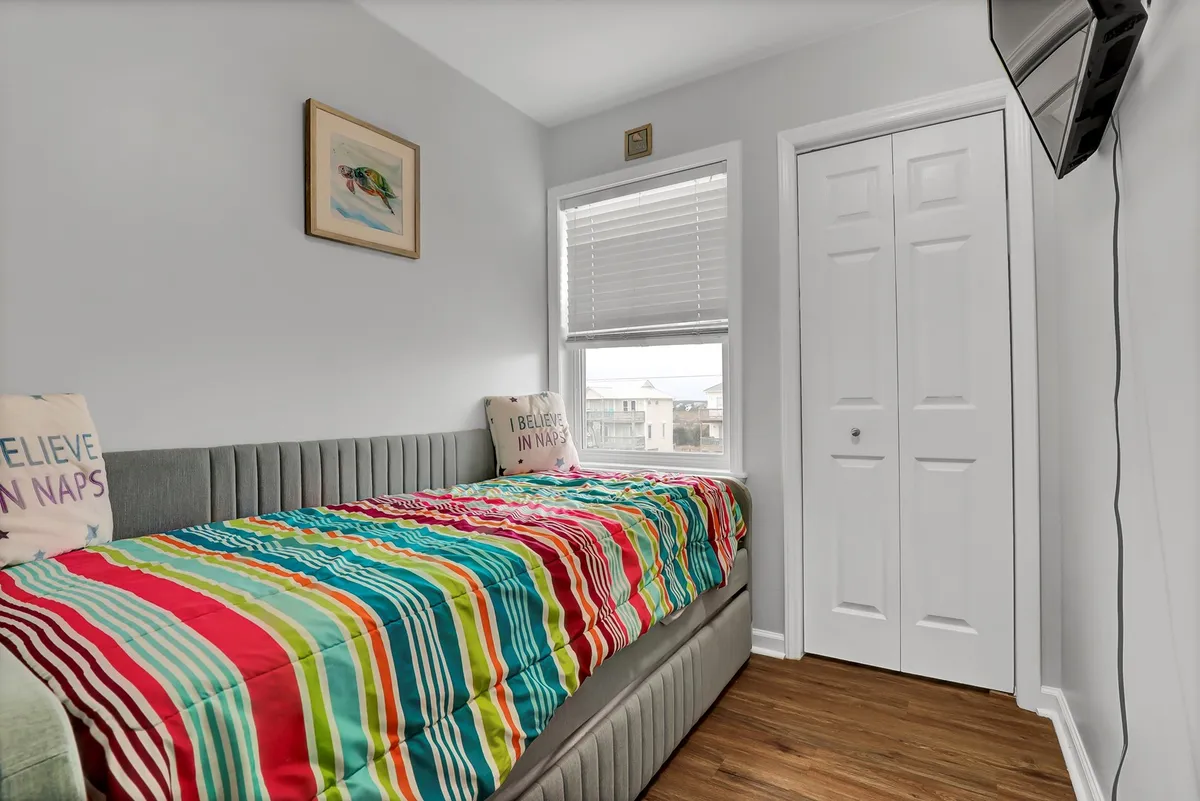 A cheerful bedroom with colorful striped bedding, wall art, a cozy bed, and natural light streaming through the window. The space feels bright and inviting, suitable for children or friends to relax and play.