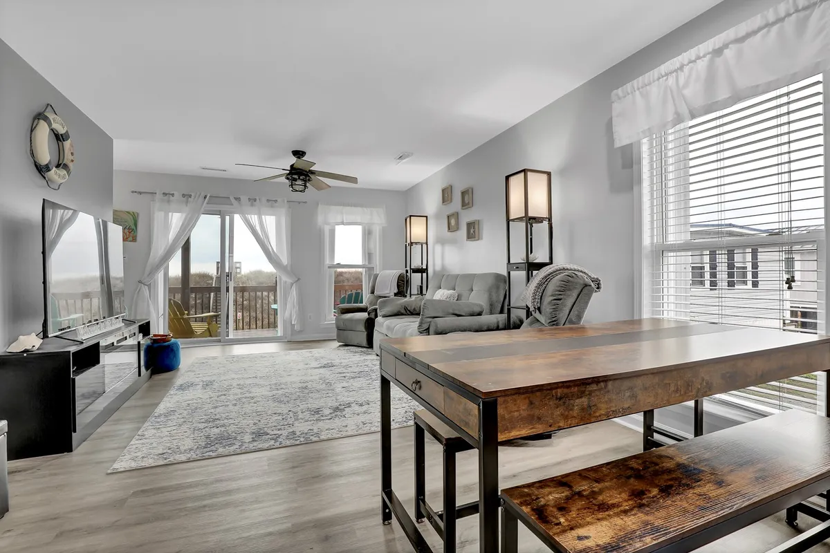A comfortable living room with a cozy seating area and a dining table set near large windows. The room is bathed in natural light, and decorative wall art adds a welcoming touch. The expansive windows frame views of the outdoors, giving a hint of the ocean beyond.