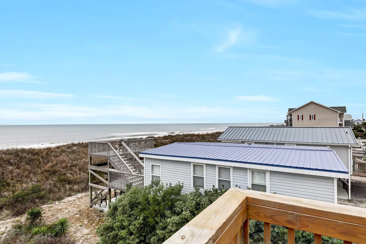 A sweeping view of the ocean from an elevated deck, with a blue roof and lush vegetation below, as well as a boardwalk leading down to the sand. The atmosphere is serene and inviting, evoking leisurely days spent by the sea and picturesque evenings watching the sunset.