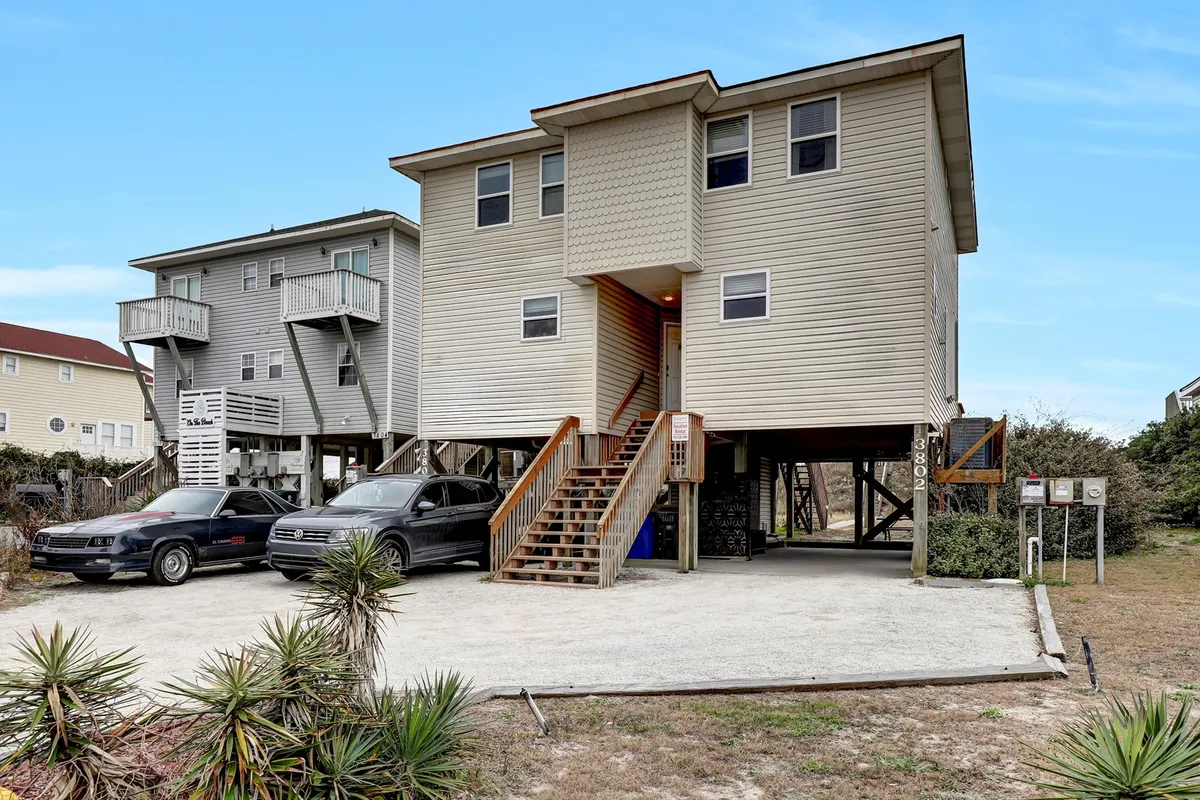A two-story duplex with a staircase entrance, ample parking, and lush surrounding vegetation. The porch offers views of the outdoors, signaling a welcoming and relaxed family environment just steps from the shore.