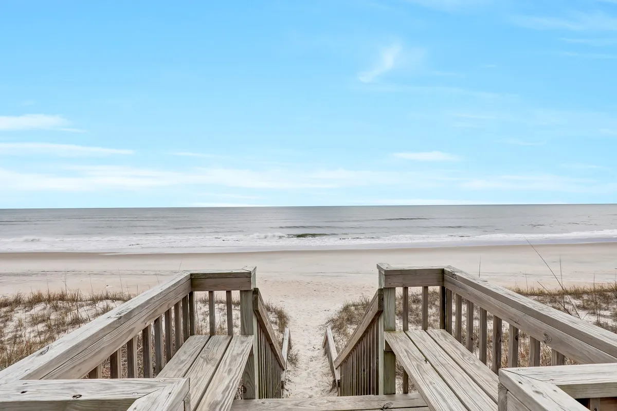 A wide view of a wooden staircase leading from the property over dune grass directly onto the sandy beach, with an expansive ocean view stretching to the horizon. The welcoming stairs invite families to transition effortlessly from the home to the shore for sun, sand, and surf.