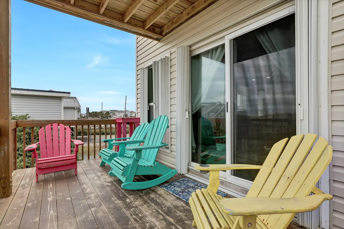 A cheerful outdoor deck with colorful Adirondack chairs positioned just outside a sliding glass door, offering inviting spots to relax. The setup is ideal for enjoying morning coffee or evening chats, with clear cues to the ocean scenery nearby.