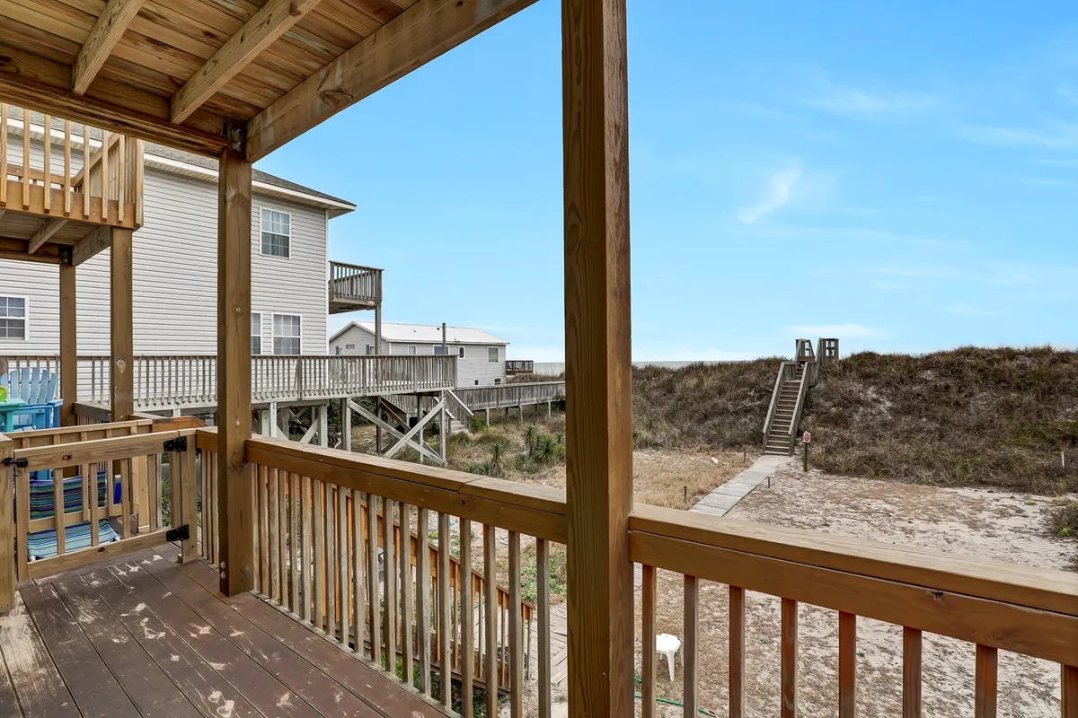 This image showcases a spacious wooden deck with a staircase leading down directly to the sandy beach. The deck is furnished for gatherings and relaxation, and the view includes neighboring properties and an inviting ocean horizon. The setting is ideal for enjoying morning coffee or evening sunsets with loved ones, with easy beach access just steps away.