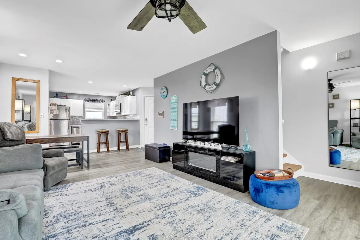 A wide-angle interior view showing the kitchen bar stools, part of the kitchen area, and a comfortable seating zone with a large TV. The scene conveys a social, open-concept living space where cooking, entertaining, and relaxing blend seamlessly.