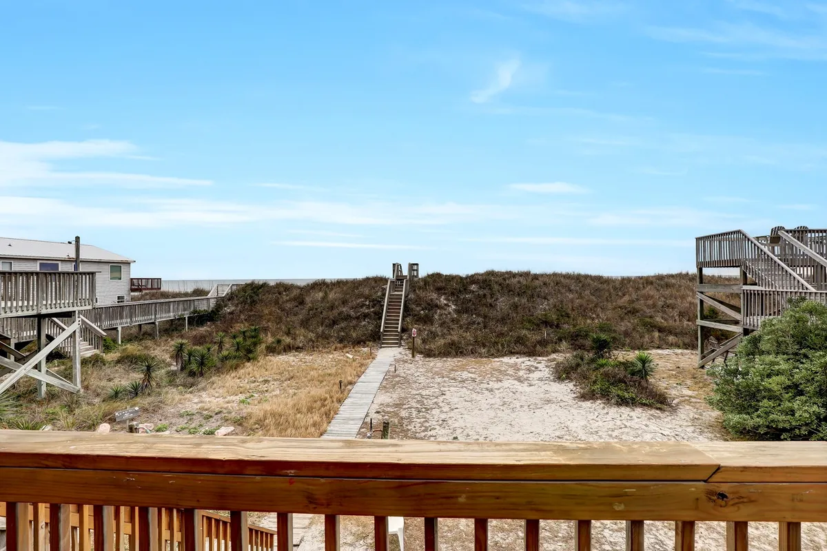 This serene view captures a wooden deck overlooking coastal dunes and an expansive ocean vista. The deck features stairs that gently descend toward the beach, surrounded by natural vegetation, evoking a sense of tranquility and direct connection with the shoreline.